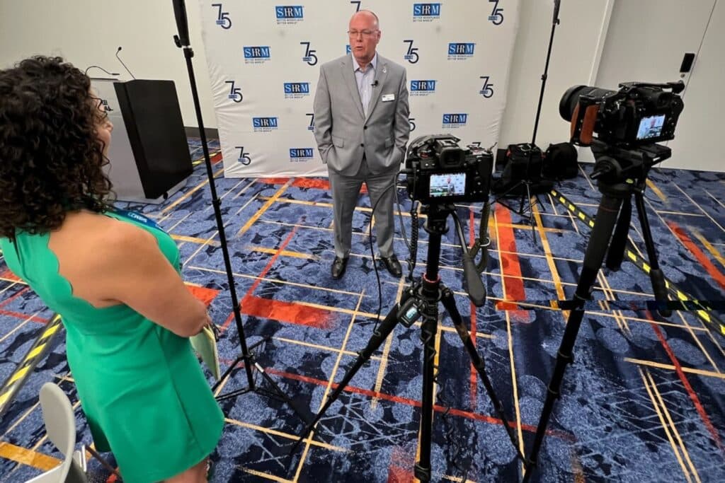 man standing behind video cameras while a woman interviews him