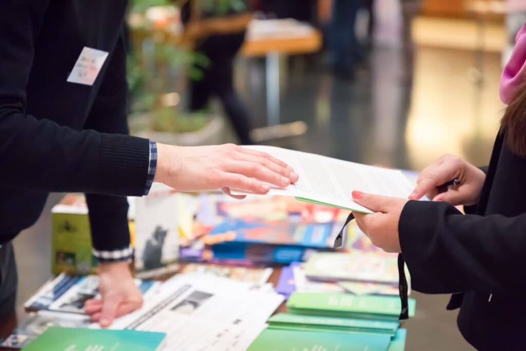 cropped photo of a person at a trade show booth holding a sheet of paper while another places their hand on the paper to explain the information